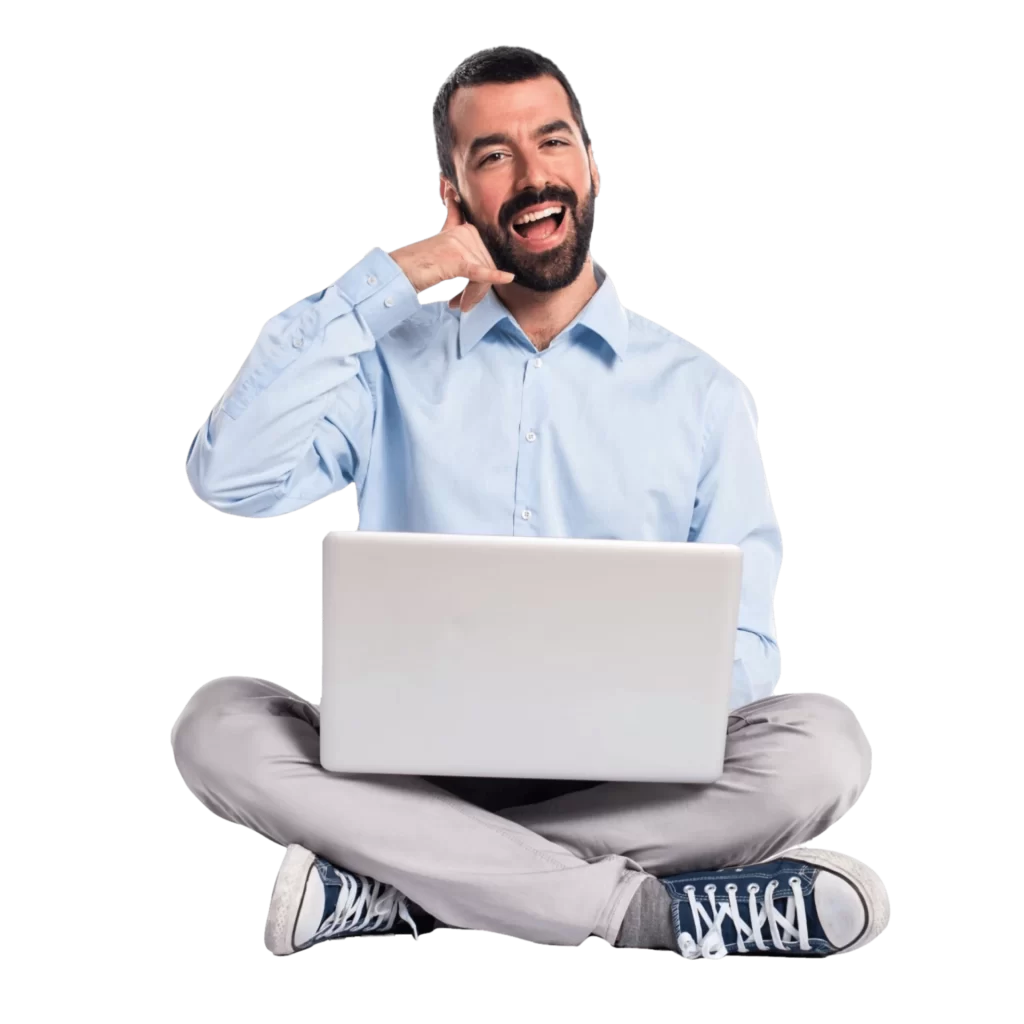 un homme souriant en chemise bleu, pantalon gris, converse bleu, assis en tailleur avec un ordinateur portable sur les genoux. Il fait le signe d'appeler par téléphone avec sa main.
